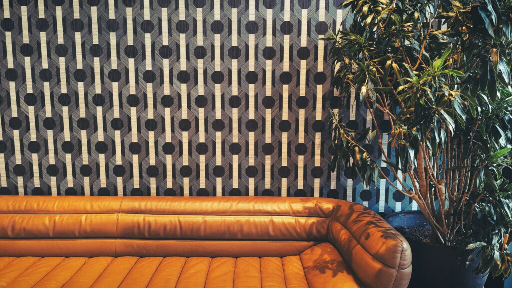 Braunes Ledersofa vor abstrakter Tapete. Rechts neben dem Sofa steht eine große Pflanze.