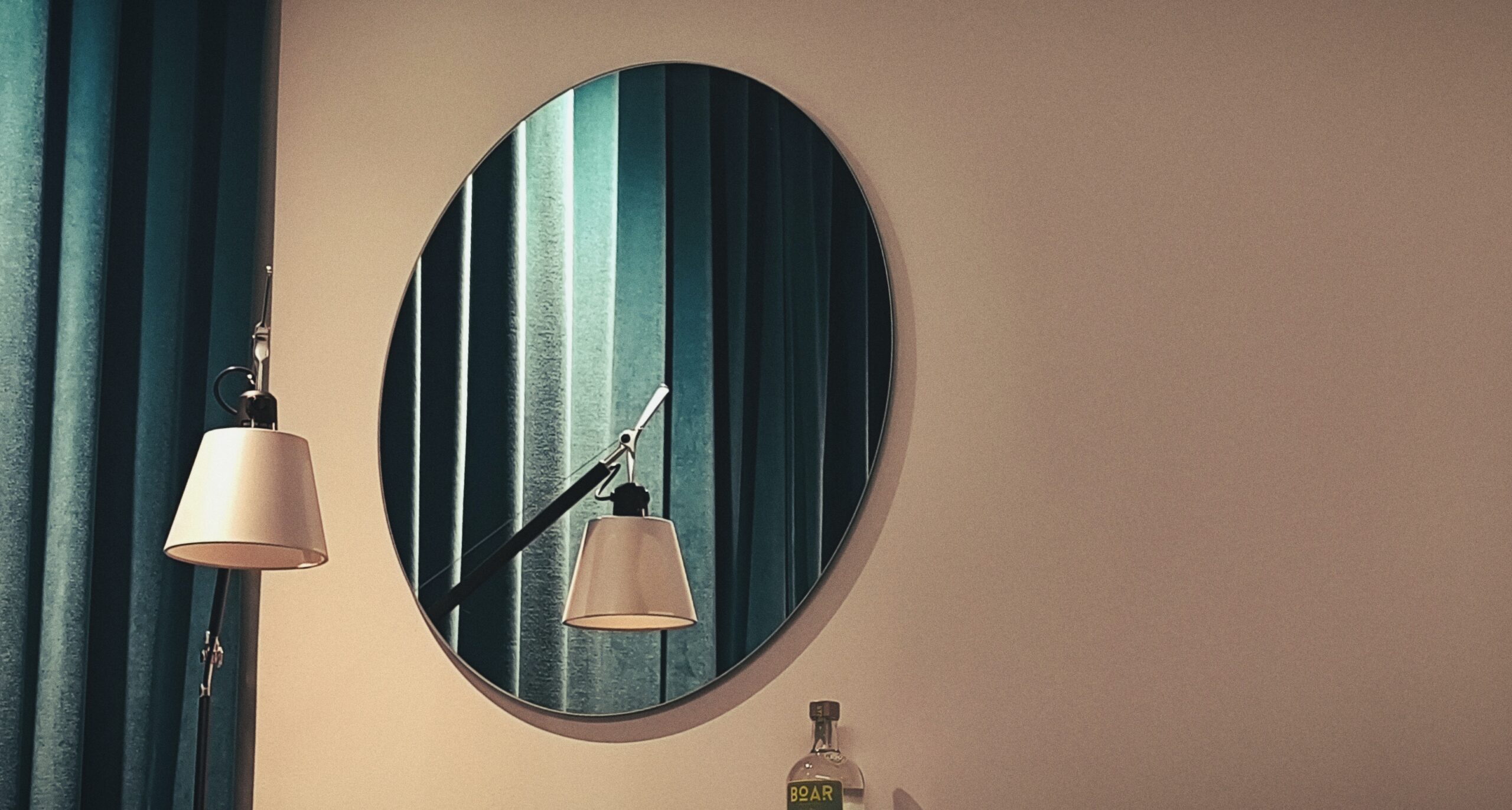 Lampe und Vorhang spiegeln sich in rundem Spiegel. Der Spiegel hängt an einer ansonsten weißen Wand.